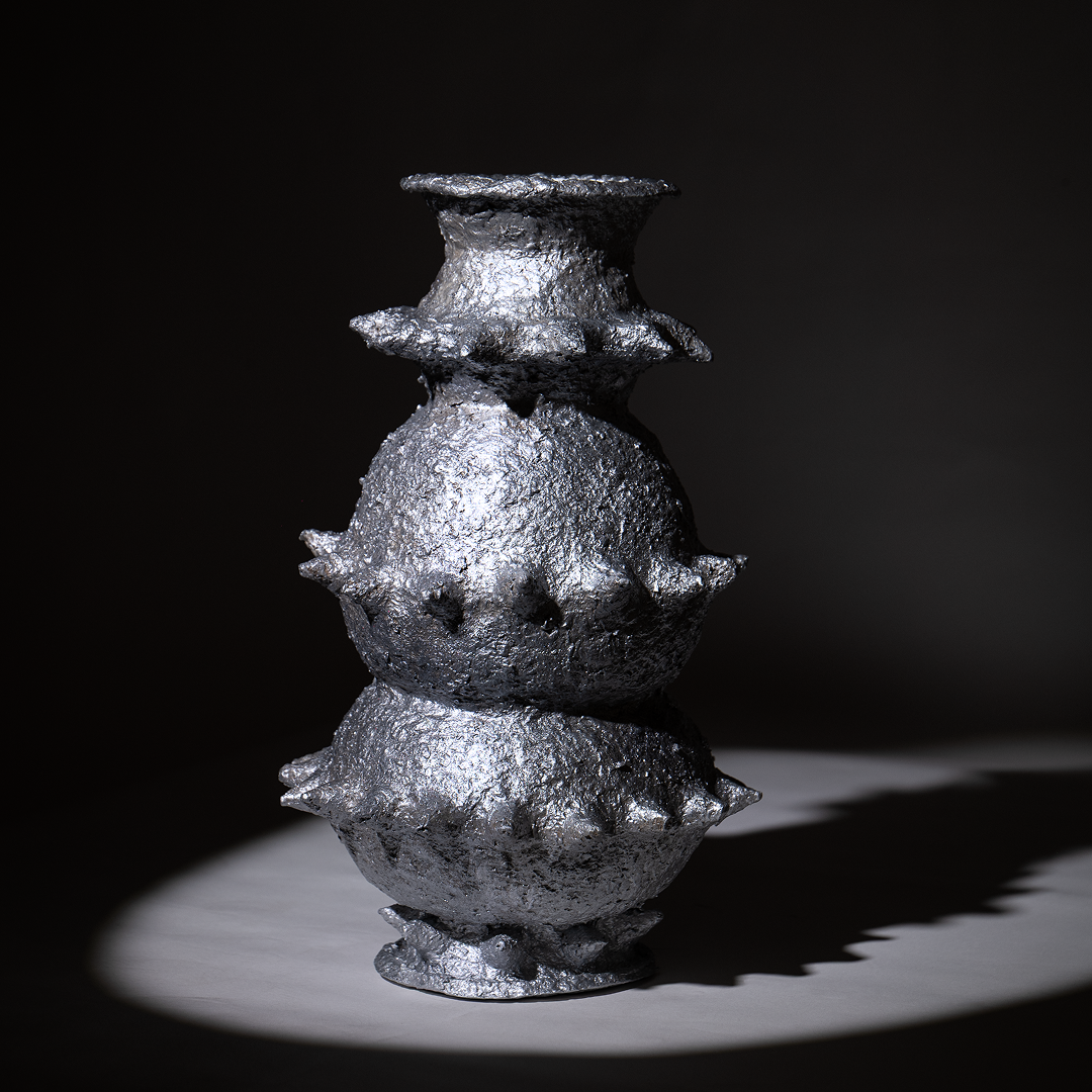 Handbuilt papier-mâché sculptural vessel “Tálä” by Pulp Fictitious, featuring stacked rounded forms and delicate spikes in a silver finish against a dark background.