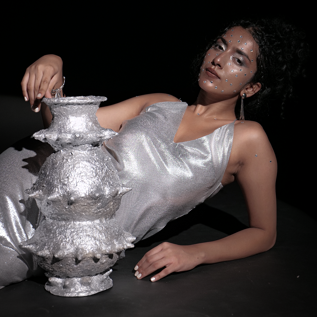 Person reclining with the handbuilt papier-mâché sculptural vessel “Tálä” by Pulp Fictitious, finished in silver, against a dark background.