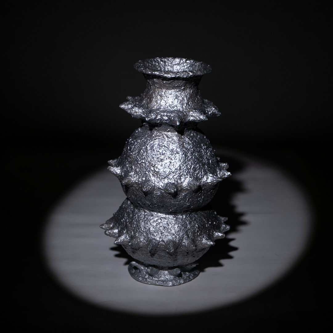 Handbuilt papier-mâché sculptural vessel “Tálä” by Pulp Fictitious, featuring stacked rounded forms and spiked details in a silver finish on a dark background. 